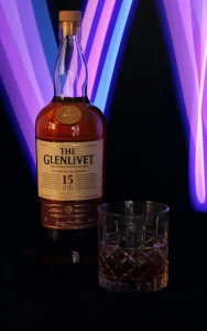 A bottle of The Glenlivet 15 Year Old Single Malt Scotch Whisky with a crystal glass, set against a vibrant neon-lit background.