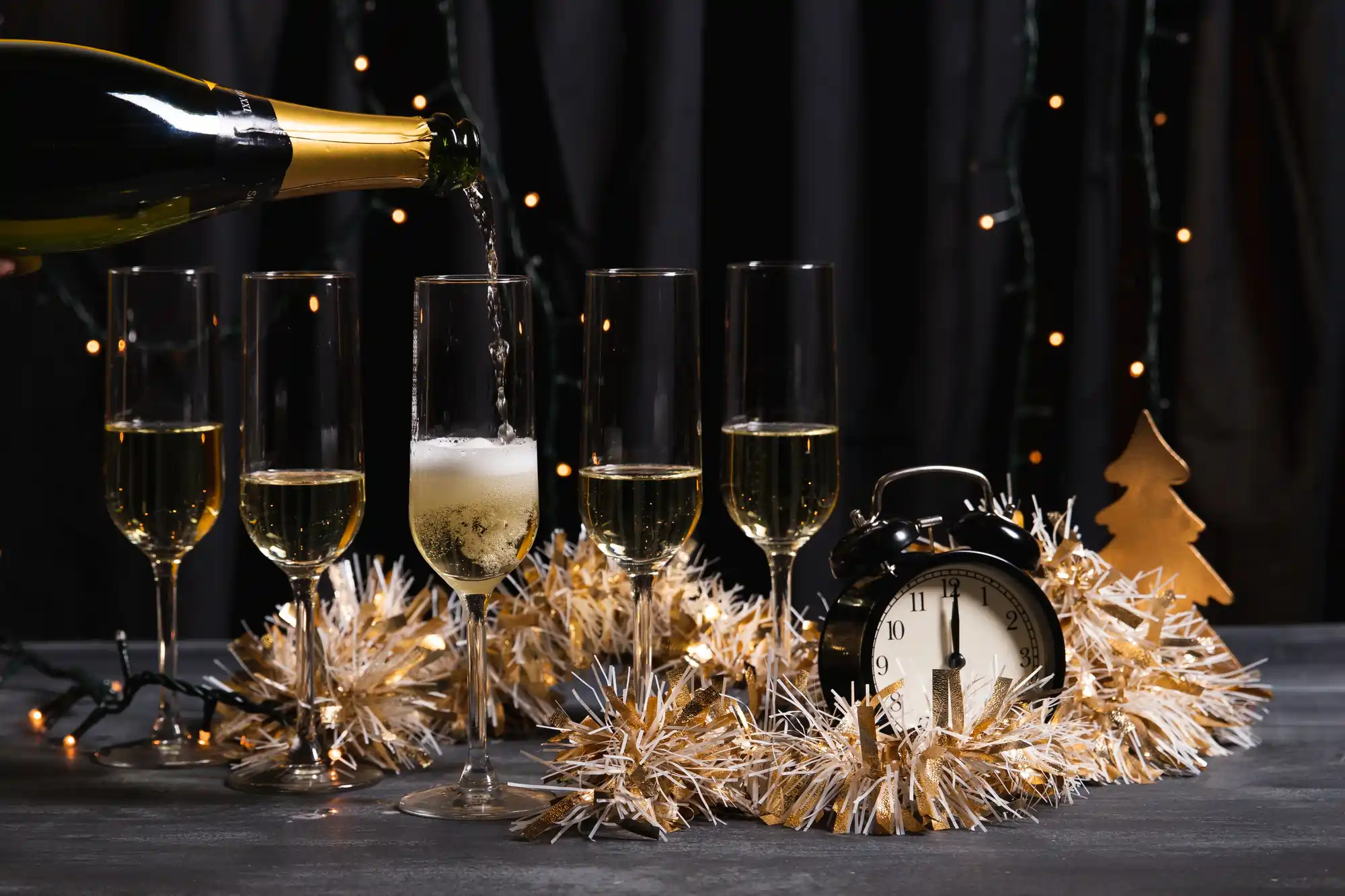 Champagne being poured into a row of glasses with a festive New Year’s Eve setup including a clock and decorations